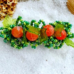 May include: A beaded hair clip with green beads and three orange agate beads. The beads are arranged in a cluster and surrounded by green leaves.
