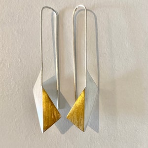 Könnte beinhalten: Ein Paar moderne geometrische Ohrringe. Jeder Ohrring hat einen langen, silberfarbenen Haken und einen rautenförmigen Anhänger. Die untere Hälfte jedes Anhängers ist goldfarben, was einen auffälligen Kontrast zum Silber bildet.