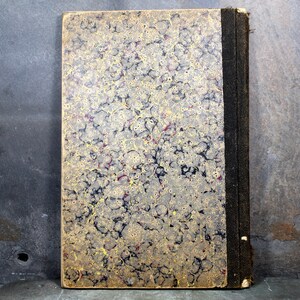 May include: Antique book with a marbled cover in shades of gold, black, and red. The spine is black with a textured appearance. The book's edges show signs of wear, suggesting its age. The cover has a unique, swirling pattern.