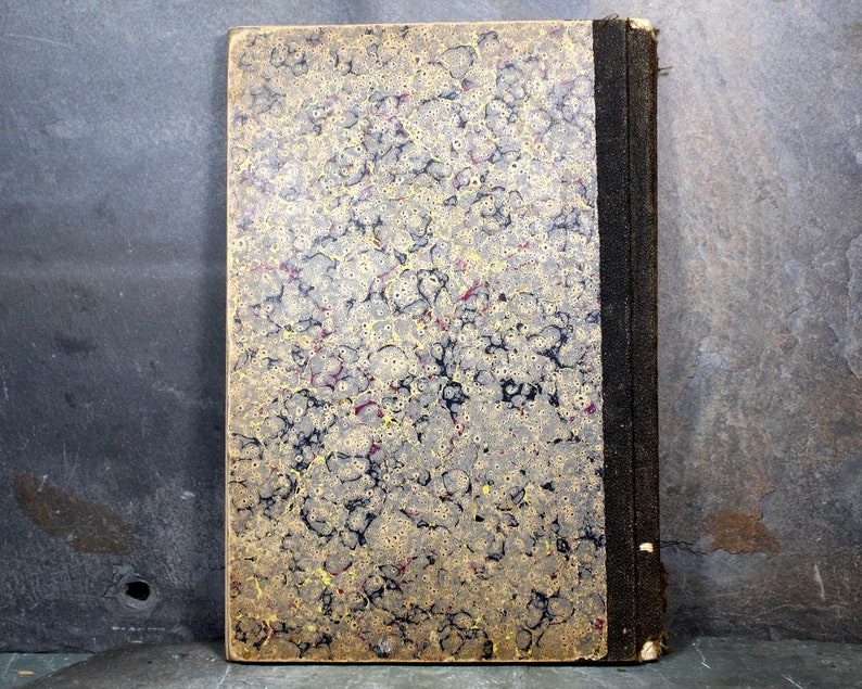 May include: Antique book with a marbled cover in shades of gold, black, and red. The spine is black with a textured appearance. The book's edges show signs of wear, suggesting its age. The cover has a unique, swirling pattern.
