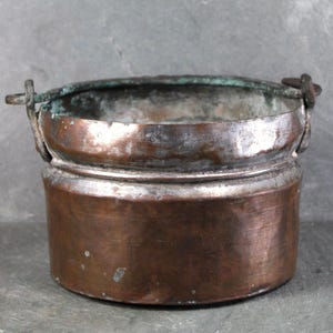 May include: A small, antique copper bucket with a handle. The bucket has a cylindrical body with a slightly flared rim and a dark patina. The handle is made of a darker metal and is attached to the sides of the bucket. The copper has a warm, reddish-brown color.