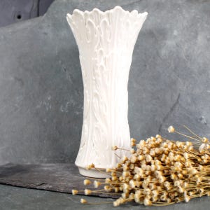 May include: A white ceramic vase with an ornate, embossed design. The vase has a wide mouth and a narrow base.
