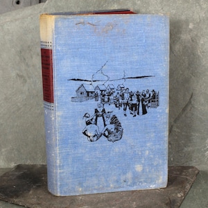 May include: A blue hardcover book with a black and white illustration of a group of people gathered around a table with turkeys on it. The book title is "The First Thanksgiving Story" and the author is "Ingri and Edgar Parin d'Aulaire".