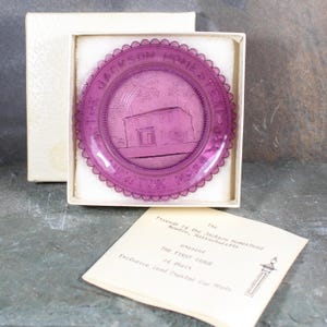 May include: A purple glass plate with a scalloped edge. The plate features an etched image of a house with the text "The Jackson Homestead 1730" around the edge.