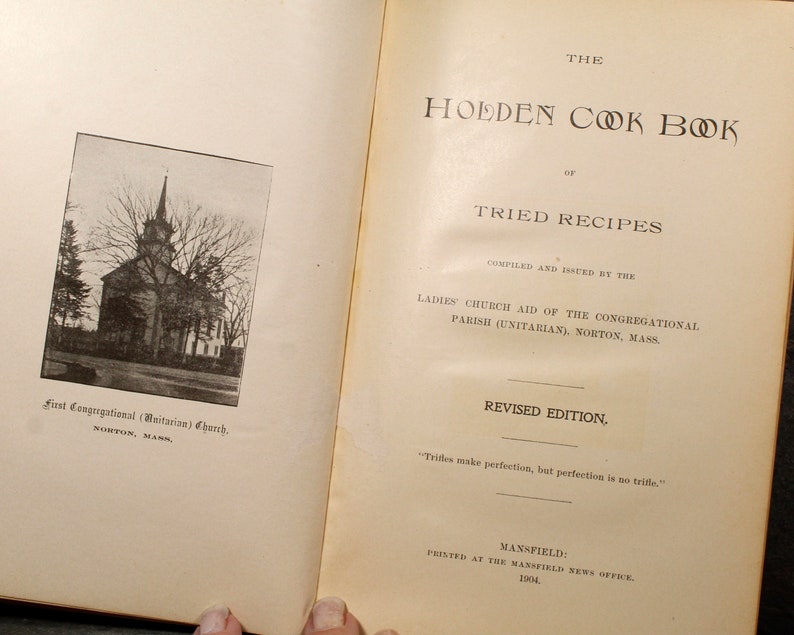 May include: Open book with the title "The Holden Cook Book of Tried Recipes." The cover features a black and white photo of a church. Text includes "Compiled and Issued by the Ladies' Church Aid" and "Revised Edition."