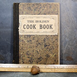 May include: An antique cookbook titled "THE HOLDEN COOK BOOK" with a mottled brown and gold cover. The book has a black spine and a cream-colored label with black text. A wooden ruler and two acorns are in the foreground.