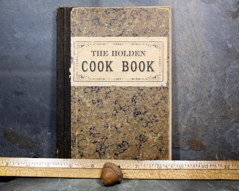 May include: An antique cookbook titled "THE HOLDEN COOK BOOK" with a mottled brown and gold cover. The book has a black spine and a cream-colored label with black text. A wooden ruler and two acorns are in the foreground.
