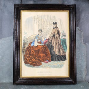 May include: A framed print of a fashion illustration from La Mode Illustrée, a French fashion magazine. The illustration depicts two women in elaborate 19th-century clothing. One woman wears a brown dress with a blue and gold jacket, while the other wears a black dress with a patterned brown shawl.