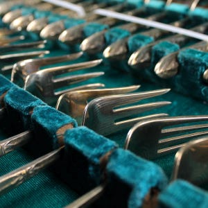 May include: A close-up of a set of silver forks in a teal velvet case. The forks are arranged in rows, with the tines facing up.