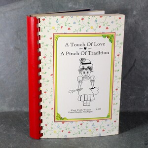 May include: A white cookbook with a red spine and a floral pattern on the cover. The title is "A Touch of Love A Pinch of Tradition" with a small heart symbol between the titles. There is a black and white illustration of a woman holding a spoon and a pot. The text at the bottom of the cover reads "B'nai B'rith Women Grand Rapids, Michigan #473".