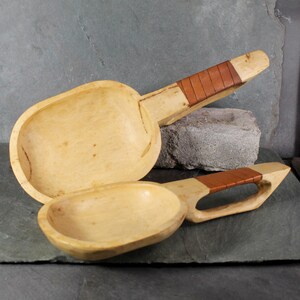 Puede incluir: Dos cucharas de servir de madera con mangos envueltos en cuero marrón claro. Las cucharas tienen una veta de madera natural y un acabado liso y pulido. Las cucharas son de diferentes tamaños, una más grande que la otra.