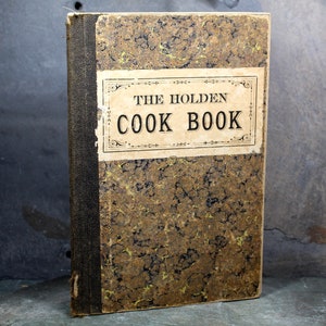 May include: An antique cookbook titled "THE HOLDEN COOK BOOK" with a worn, patterned cover in shades of brown and gold. The spine is dark, and the title is displayed on a rectangular label. The book appears aged and well-loved.