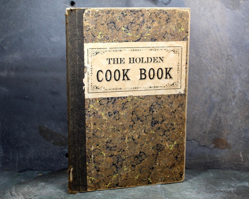 May include: An antique cookbook titled "THE HOLDEN COOK BOOK" with a worn, patterned cover in shades of brown and gold. The spine is dark, and the title is displayed on a rectangular label. The book appears aged and well-loved.