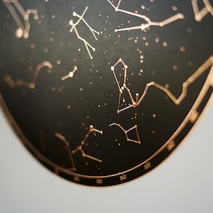 K&ouml;nnte beinhalten: Eine schwarze, runde Wanddekoration mit einem kupferfarbenen Design von Sternbildern. Die Sternbilder sind mit kleinen Punkten und Linien umrandet.