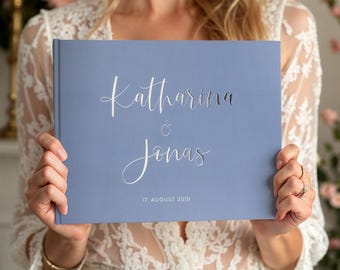May include: A light blue wedding guest book with silver lettering. The book says "Katharina & Jonas" and "17. August 2019".