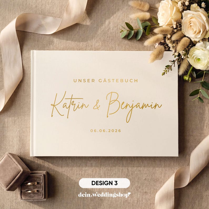 May include: A cream-colored wedding guest book with gold lettering that reads "UNSER G&Auml;STEBUCH" and the names "Katrin & Benjamin" along with the date "06.06.2026". The book is surrounded by a bouquet of flowers, a ribbon, and two ring boxes.