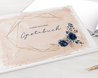 May include: A white guest book with a blush pink watercolor background and gold geometric frame. The text "UNSERE HOCHZEIT Gästebuch" is written in navy blue cursive. The cover is decorated with blue watercolor flowers.