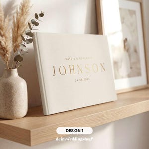 May include: A cream-colored wedding guest book with gold lettering that reads "JOHNSON" and the date "24.08.2024". The names "KATRIN & BENJAMIN" are above the last name. The book is on a wooden shelf with a vase of dried flowers.
