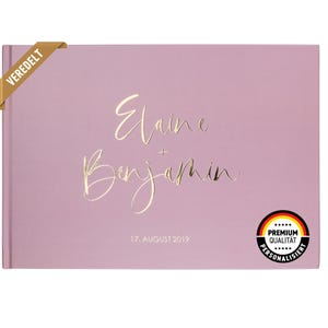 May include: A dusty rose colored guest book with gold foil lettering that reads "Elaine + Benjamin" and "17. AUGUST 2019". The book has a gold foil sticker that reads "VEREDELT" on the top left corner and a gold foil sticker that reads "PREMIUM QUALITAT PERSONALISIERT" on the bottom right corner.