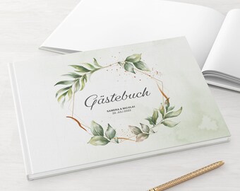 May include: A white guest book with the German word "Gästebuch" and the names "Sandra & Nicolai" and the date "22. Juli 2022" printed on the cover. The cover features a gold geometric frame with green leaves and gold accents. A gold pen rests on the surface.