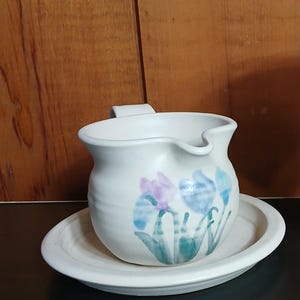 May include: A white ceramic gravy boat with a matching saucer. The boat features a floral design in blue, purple, and green. The set is placed on a dark surface against a wooden background.
