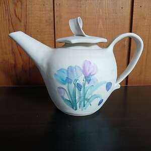 May include: Cream-colored ceramic teapot with a floral design featuring blue and purple irises. The teapot has a curved handle, a spout, and a lid with a decorative handle. The background is a wooden surface.