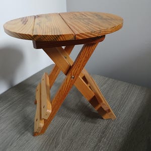 May include: A small, round, folding wooden side table. The tabletop is made of two wooden planks and has a natural wood grain finish. The legs are a crisscross design, and the table is a light brown color.