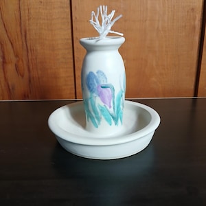 May include: A white ceramic oil lamp with a floral design in blue, purple, and green. The lamp has a small, round base and a taller, cylindrical body. A white wick is visible at the top. The lamp sits in a matching white dish.