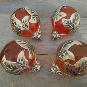May include: Four decorative cabinet knobs with a reddish-orange, rounded glass-like center. Each knob features a silver-colored metal leaf design and a screw for attachment. The knobs are arranged on a wooden surface.