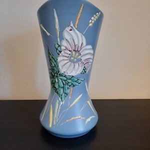 May include: A light blue ceramic vase with a flared top and a bulbous base. The vase features a white and purple floral design with green leaves and yellow and brown accents. The vase is approximately 10 inches tall.