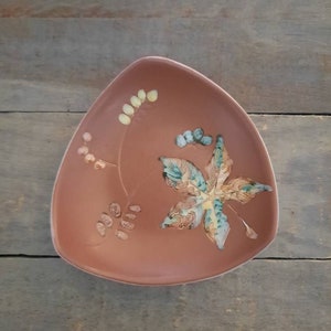 May include: A terracotta-colored, triangular ceramic plate with a raised floral design. The design features a stylized leaf in shades of blue, green, and yellow, along with smaller floral accents. The plate has a slightly raised edge.