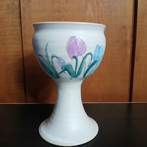May include: A white ceramic goblet with a wide bowl and a flared base. The goblet is hand-painted with purple and blue tulip flowers and green leaves. The goblet is a decorative piece.