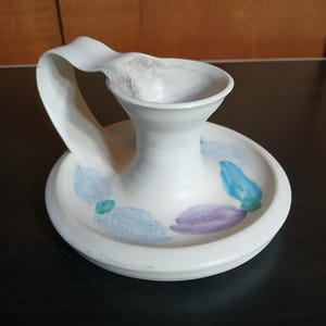 May include: A white ceramic pitcher with a wide, flat base and a curved handle. The base is decorated with blue, green, and purple abstract shapes. The pitcher has a unique, organic design.