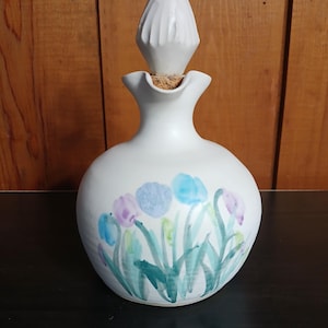May include: White ceramic bottle with a bulbous body and a tall, pointed stopper. The bottle is decorated with hand-painted blue, purple, and green floral designs. The bottle has a cork stopper.