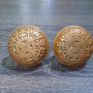May include: A pair of antique bronze door knobs, each featuring detailed floral and geometric patterns. The knobs are spherical and display a warm, aged patina. They are set against a dark blue wooden surface, highlighting their intricate design.