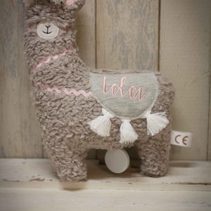 May include: A gray plush llama toy with a pink embroidered name "Lola" on its back. The llama has white tassels and a white rolling wheel on the bottom.
