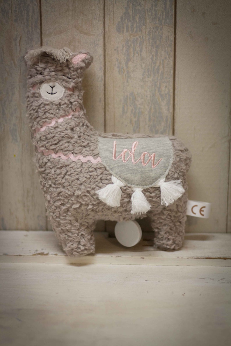 May include: A gray plush llama toy with a pink embroidered name "Lola" on its back. The llama has white tassels and a white rolling wheel on the bottom.