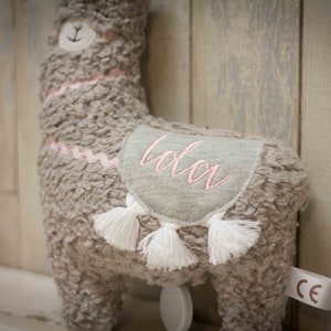 May include: A gray plush llama toy with a pink embroidered name "Lola" on its back. The llama has white tassels and a pink ribbon around its neck.
