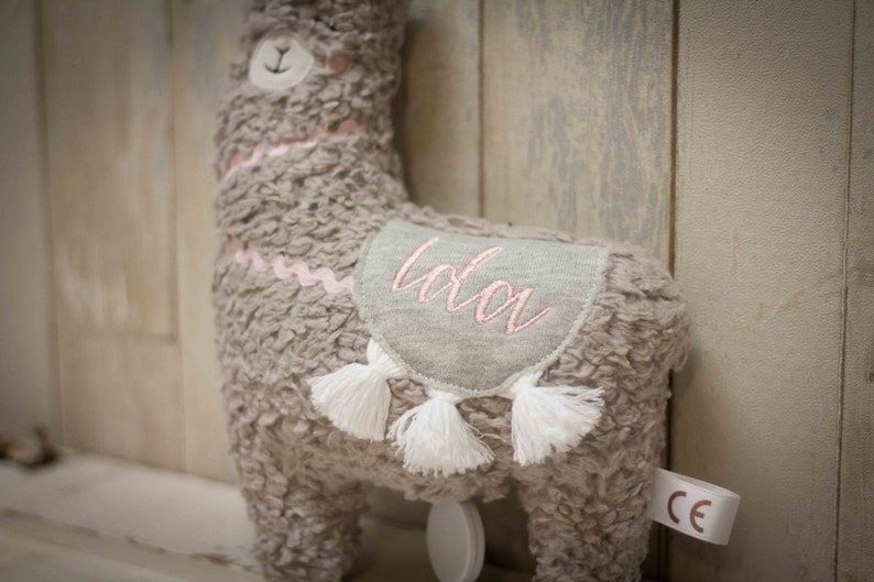 May include: A gray plush llama toy with a pink embroidered name "Lola" on its back. The llama has white tassels and a pink ribbon around its neck.