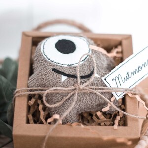 May include: A brown plush monster toy with one eye and a toothy grin, tied with twine and placed in a brown cardboard box with shredded paper. The box has a white tag attached that says "Mutmonster".