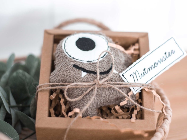 May include: A brown plush monster toy with one eye and a toothy grin, tied with twine and placed in a brown cardboard box with shredded paper. The box has a white tag attached that says "Mutmonster".