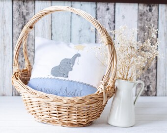 May include: A wicker basket with a blue and white pillow featuring an embroidered elephant design. The pillow has the text "Alice Marie" embroidered on it. The basket is sitting on a white surface with a white pitcher and dried flowers.