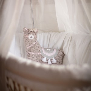 May include: A soft, gray llama-shaped pillow with pink and gray accents and white tassels sits in a white crib with a white canopy.