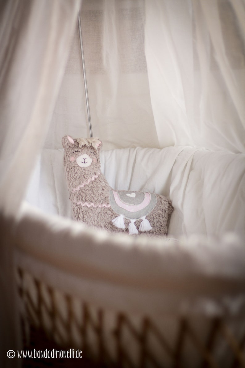 May include: A soft, gray llama-shaped pillow with pink and gray accents and white tassels sits in a white crib with a white canopy.