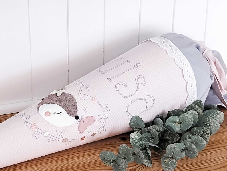 May include: Pink and gray school cone with a cute embroidered deer and butterfly design. The cone has a white lace trim and the word "Schule" embroidered on it.