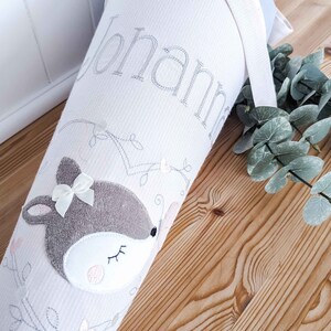 May include: A white fabric cone with a deer applique and the name "Johanna" embroidered on it. The cone is decorated with a white lace trim and a light blue ribbon.