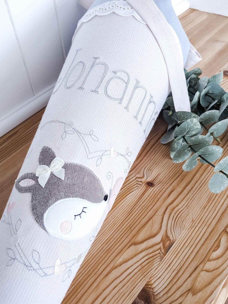May include: A white fabric cone with a deer applique and the name "Johanna" embroidered on it. The cone is decorated with a white lace trim and a light blue ribbon.