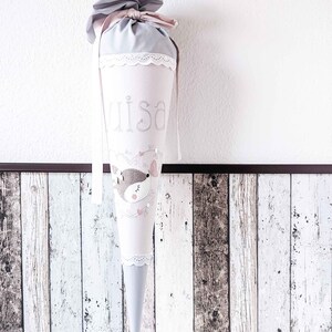 May include: A gray and pink fabric cone with a white lace trim. The cone has a pink ribbon tied around the top and the name "Lisa" embroidered on the front. A small embroidered animal with pink accents is on the front of the cone.