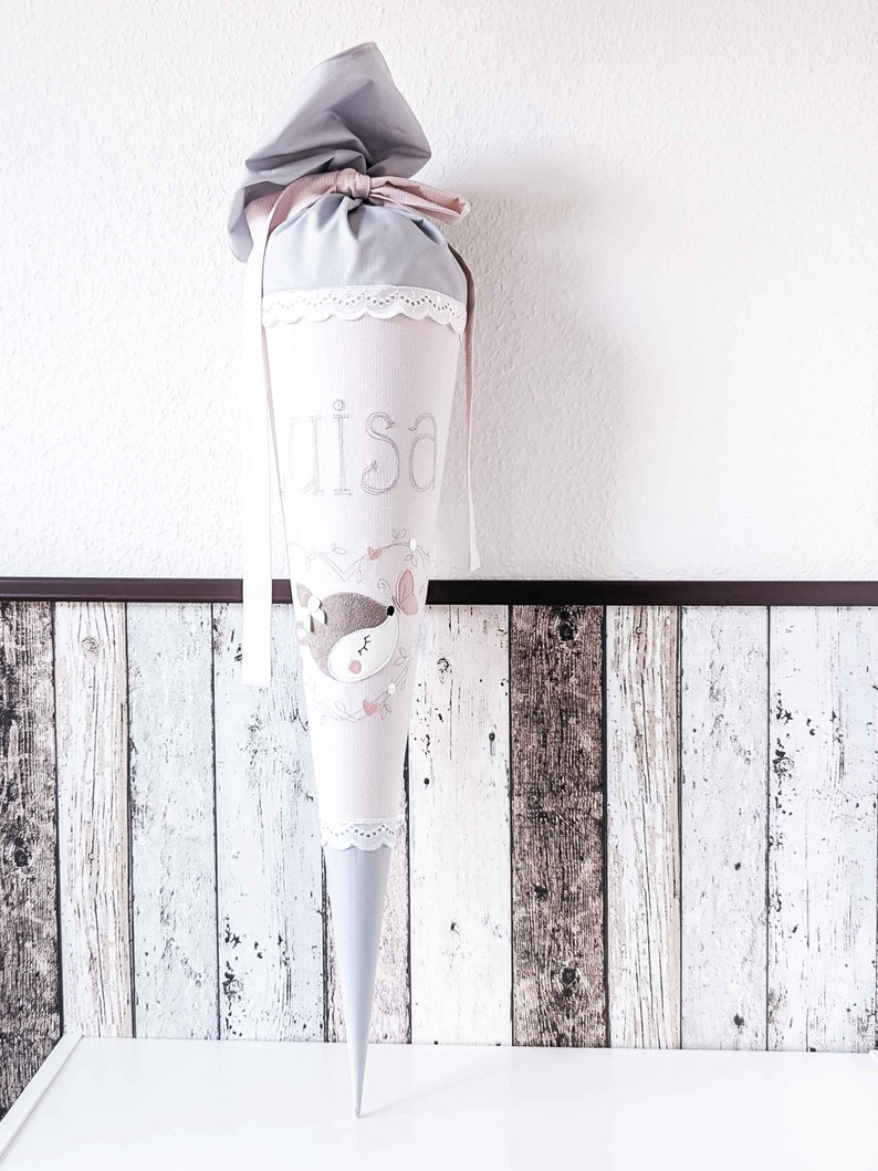 May include: A gray and pink fabric cone with a white lace trim. The cone has a pink ribbon tied around the top and the name "Lisa" embroidered on the front. A small embroidered animal with pink accents is on the front of the cone.
