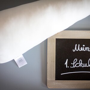 May include: A white pillow with a tag on it is next to a small chalkboard with the words "Mein 1. Schultag" written in white chalk.